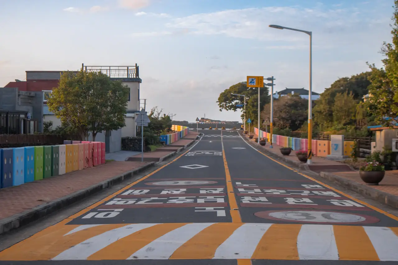 An empty two-way road on Jeju with a rainbow-coloured wall and trees beside it, yellow markings on the asphalt