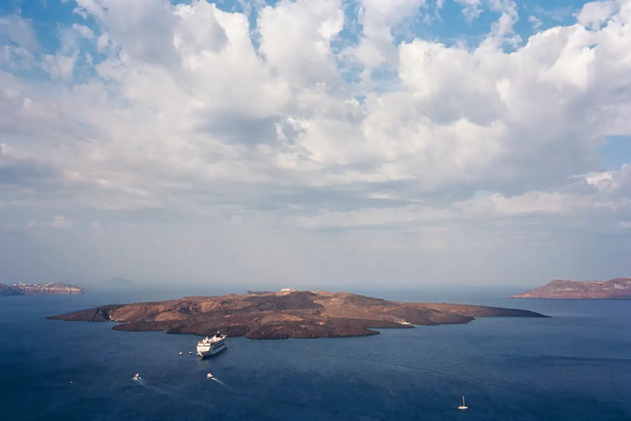 从高处俯瞰圣托里尼火山湾，深蓝海面上一艘白色邮轮停在新卡梅尼岛旁，上方是大片云层