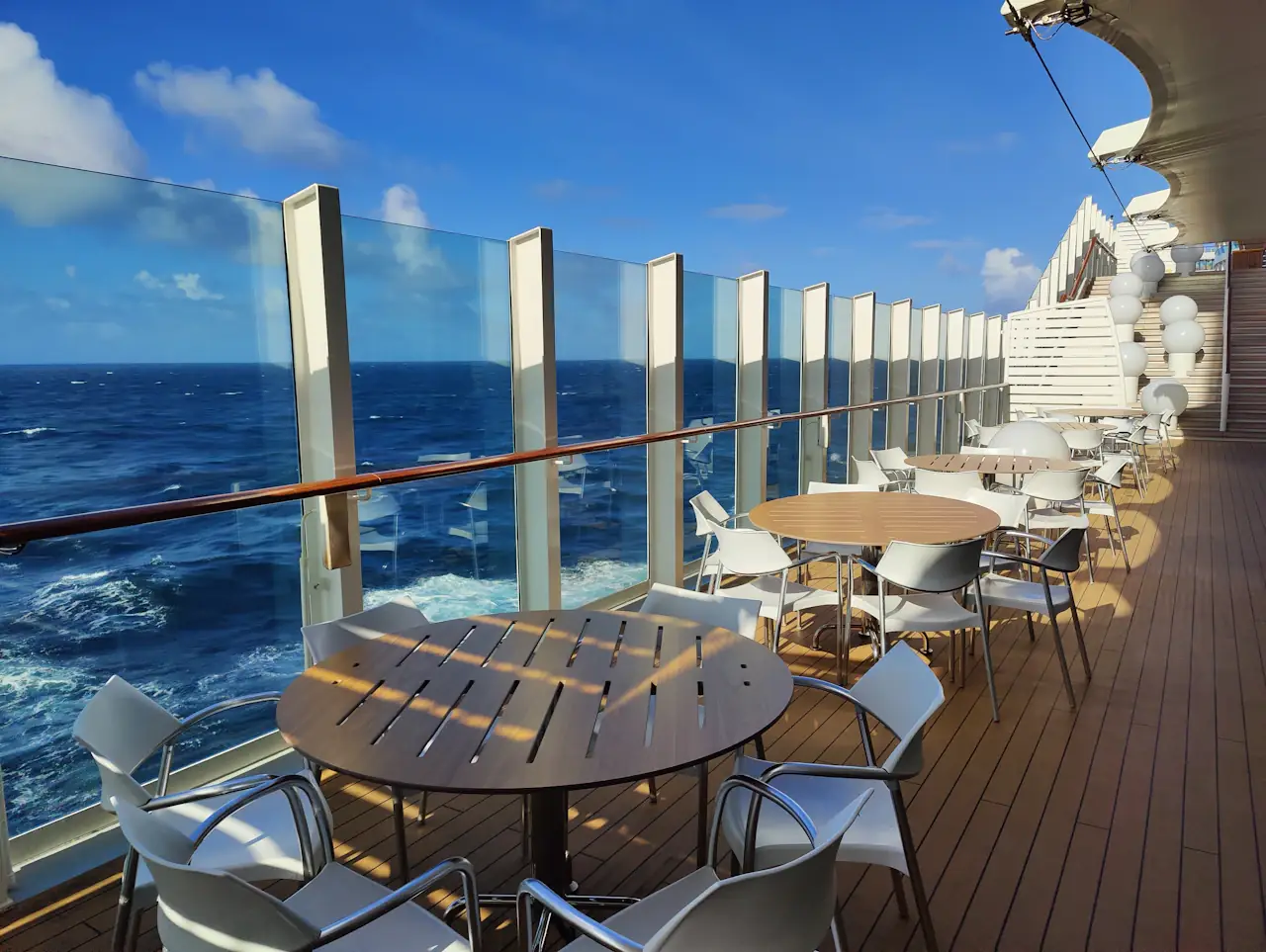 Empty round tables and white chairs on an open cruise ship deck behind glass railings, with deep blue sea and sky beyond