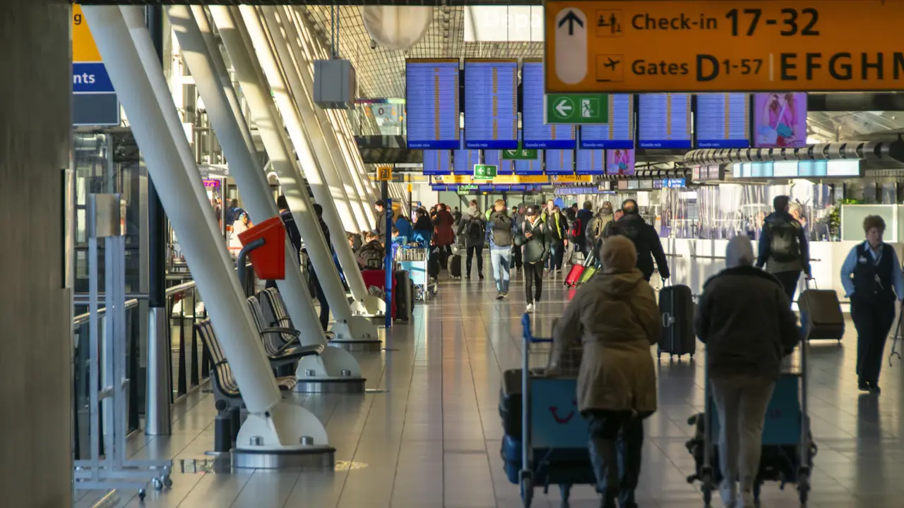 Passengers pushing luggage carts through the check-in hall at Amsterdam Schiphol Airport, departure screens overhead