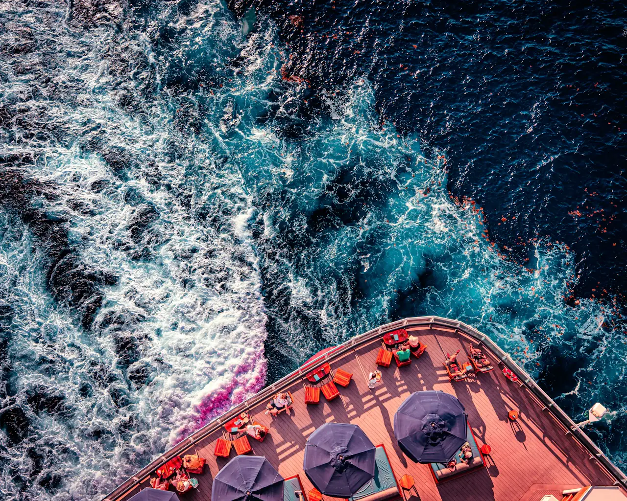 Aerial view of a cruise ship's rear deck with umbrellas and loungers beside the churning white wake