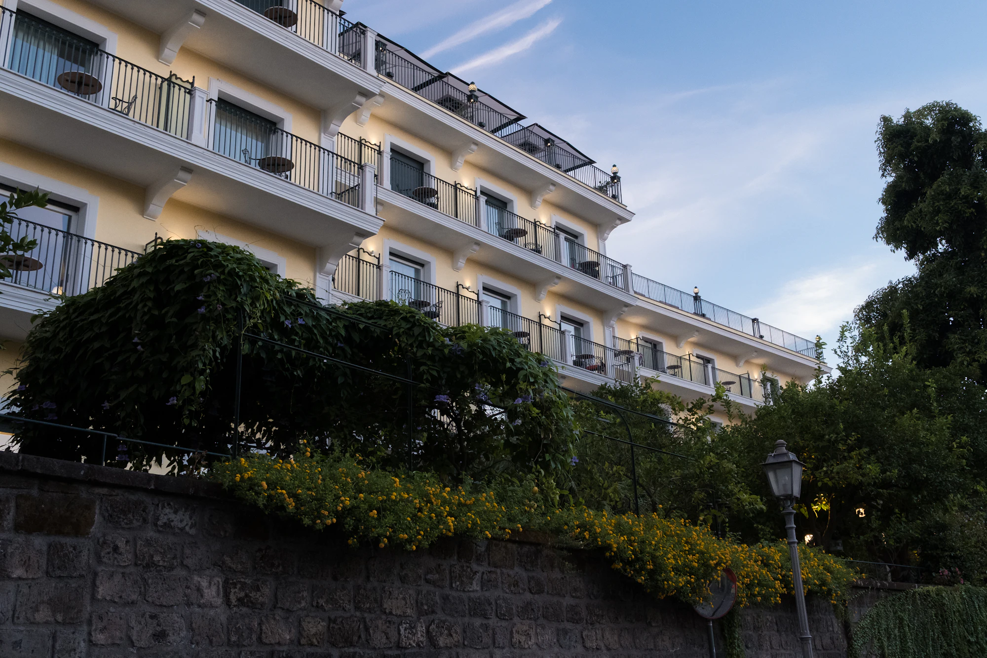Evening light on a white hotel facade with rows of balconies above a stone wall and trees