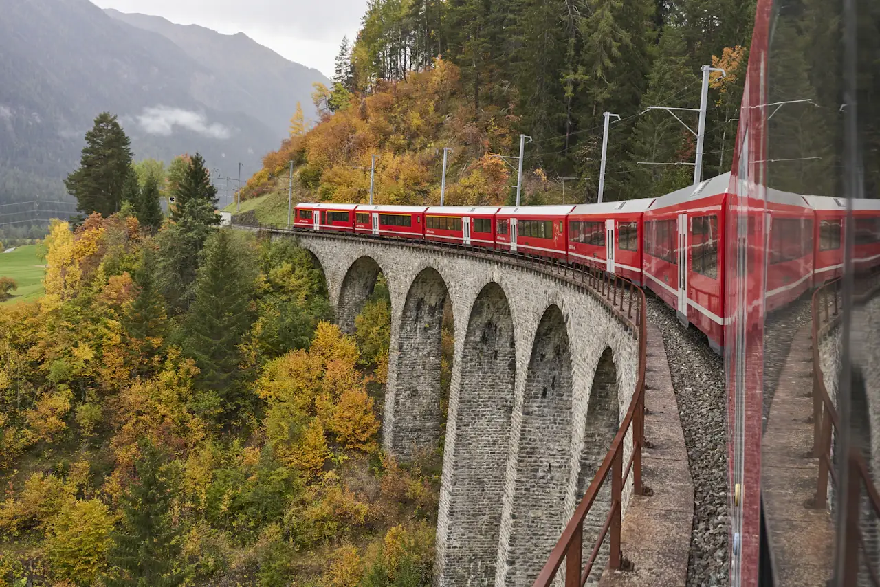 A red Rhaetian Railway train crossing a stone viaduct on the Albula line in autumn, golden larch forest on the slope