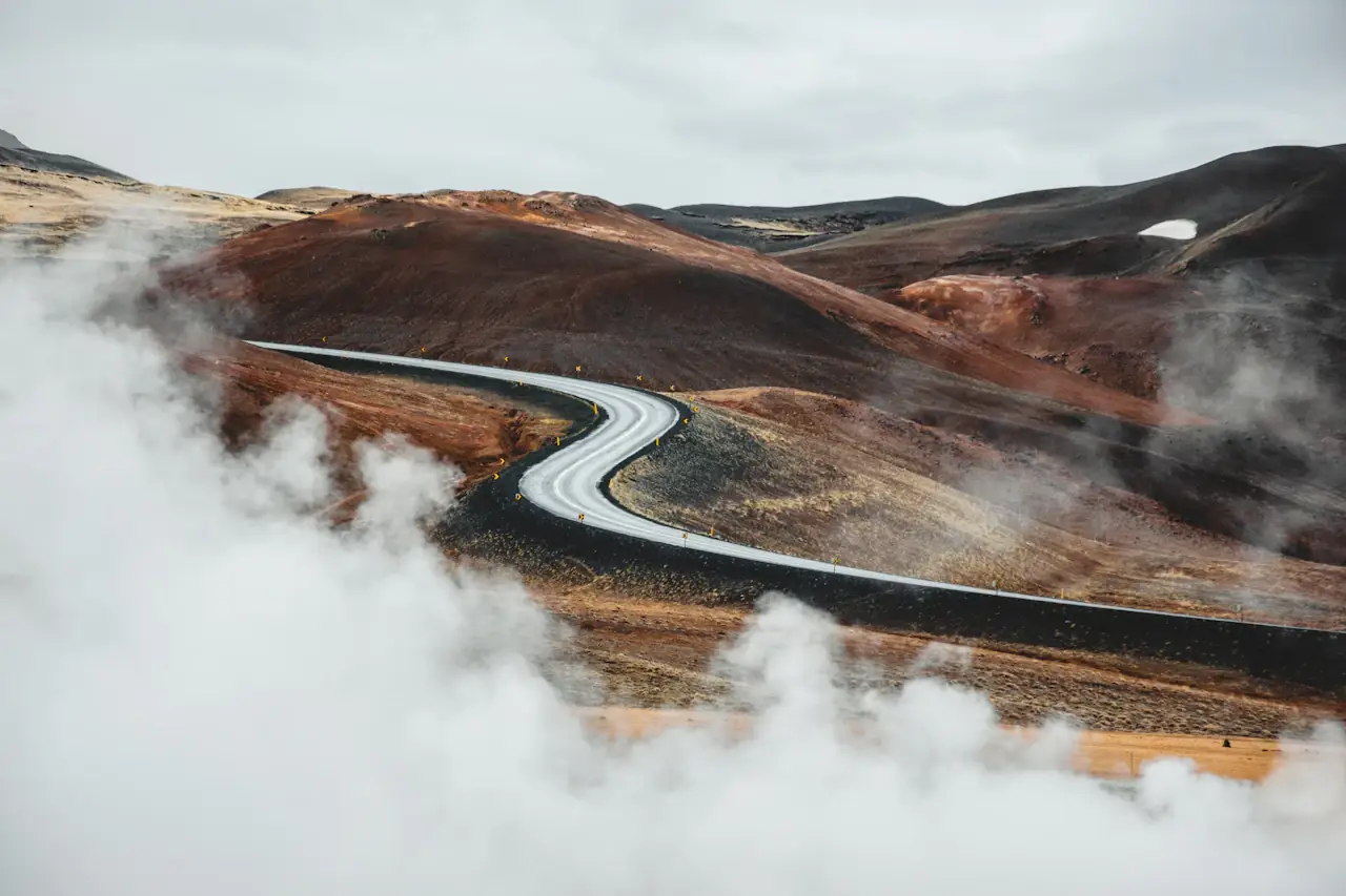 An empty winding road through steaming red-brown hills in the Mývatn geothermal area, Iceland