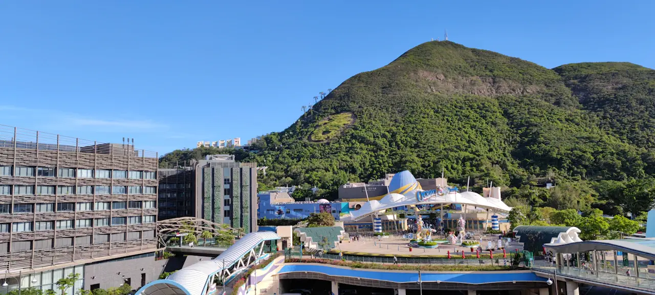 The Ocean Park entrance area in Hong Kong: white tent-roofed structures and glass buildings at the foot of a green hill under a blue sky