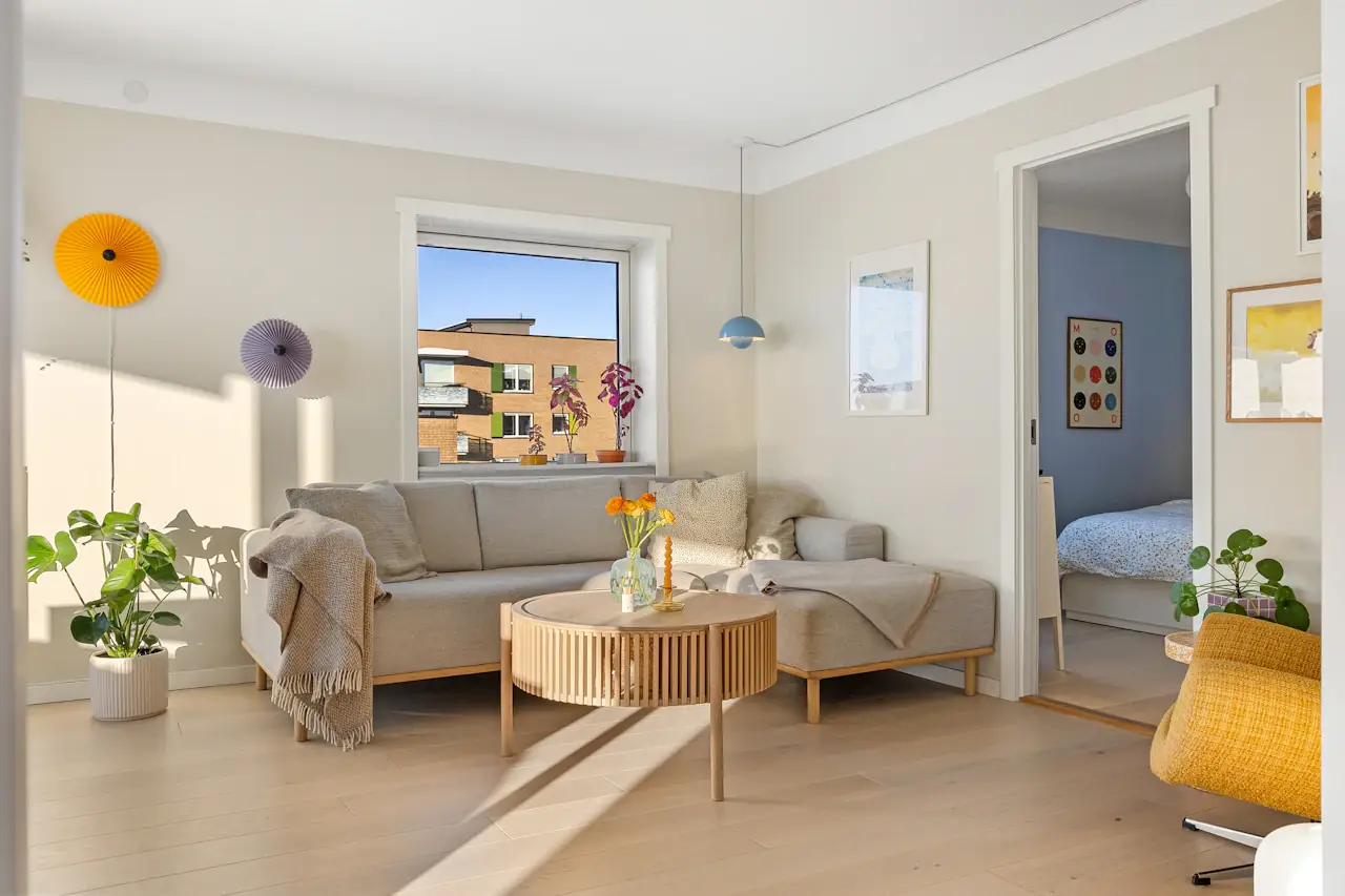 A bright apartment living room with a pale sofa, a round wooden coffee table and a window, a bedroom door beyond